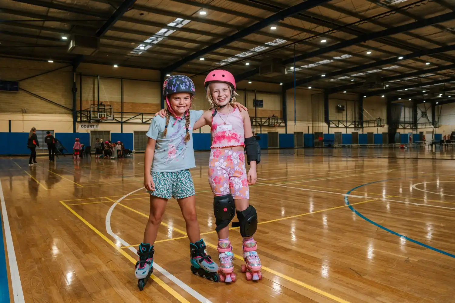 Two smiling young girls wearing helmets, knee pads, and roller skates stand arm-in-arm on a large indoor timber sports court with a high ceiling.